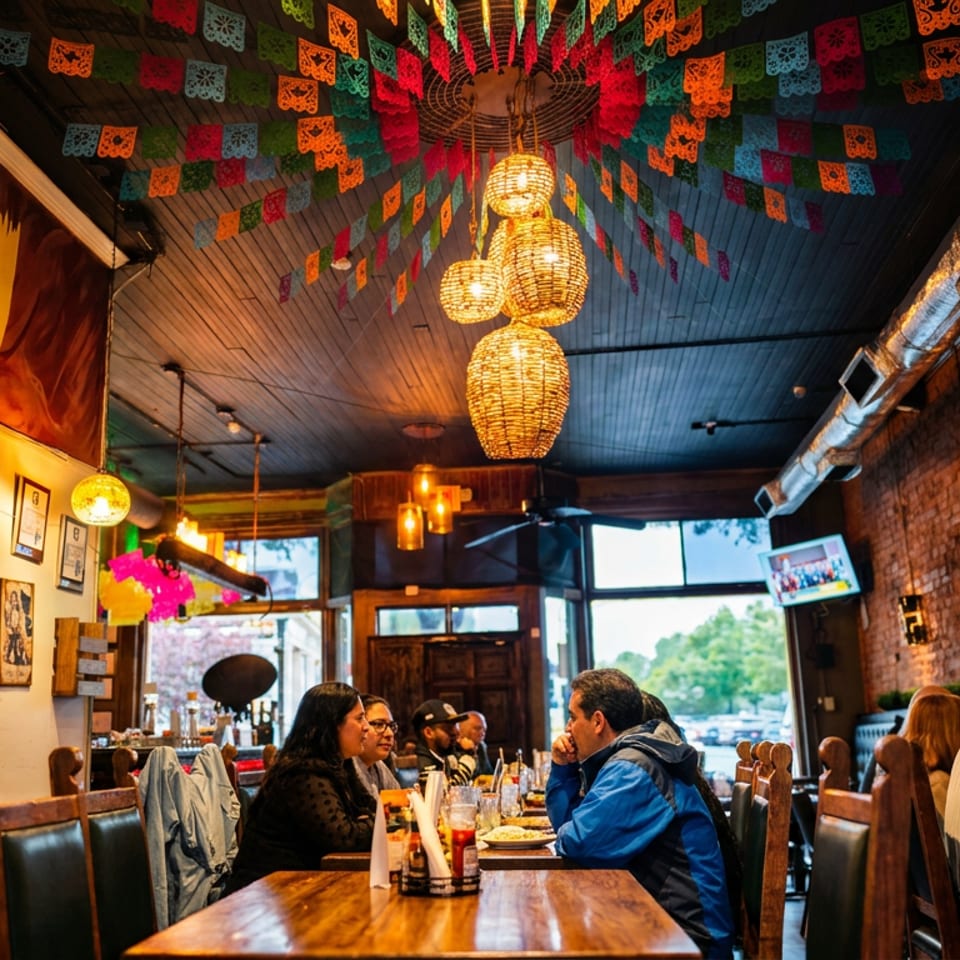 Bustling restaurant dining room with colorful papel picado banners, woven pendant lights, exposed brick walls, and diners at wooden tables.