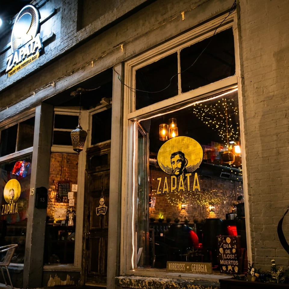 Nighttime exterior of Zapata restaurant with illuminated signage, glowing windows, outdoor seating, and festive string lights.