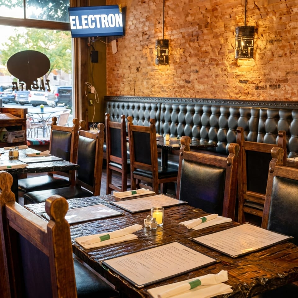 Rustic restaurant dining room with wooden tables, menus, candles, leather chairs, tufted banquette, exposed brick walls, and a storefront window.