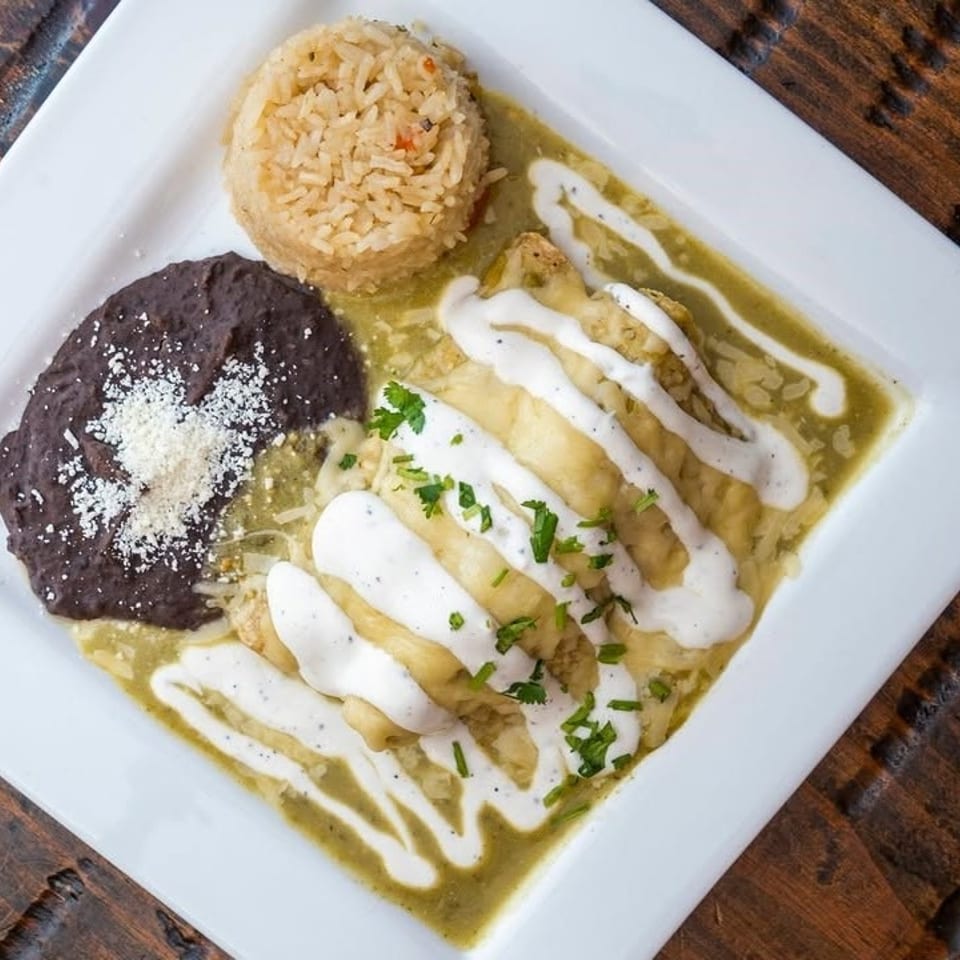 Plate of enchiladas topped with cream and cilantro, served with Mexican rice and refried black beans in green sauce.