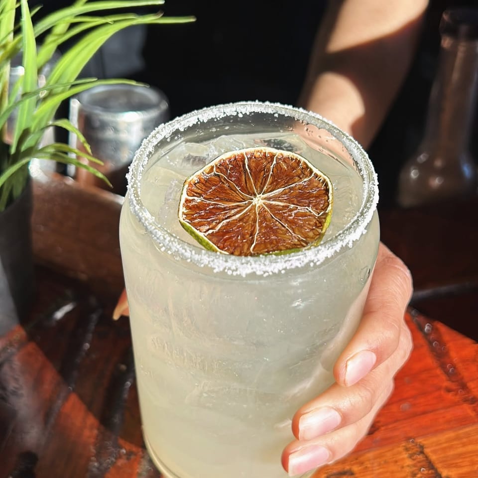 Icy pale cocktail with a salt-rimmed glass, dried lime garnish, and a hand holding it over a wooden restaurant table.