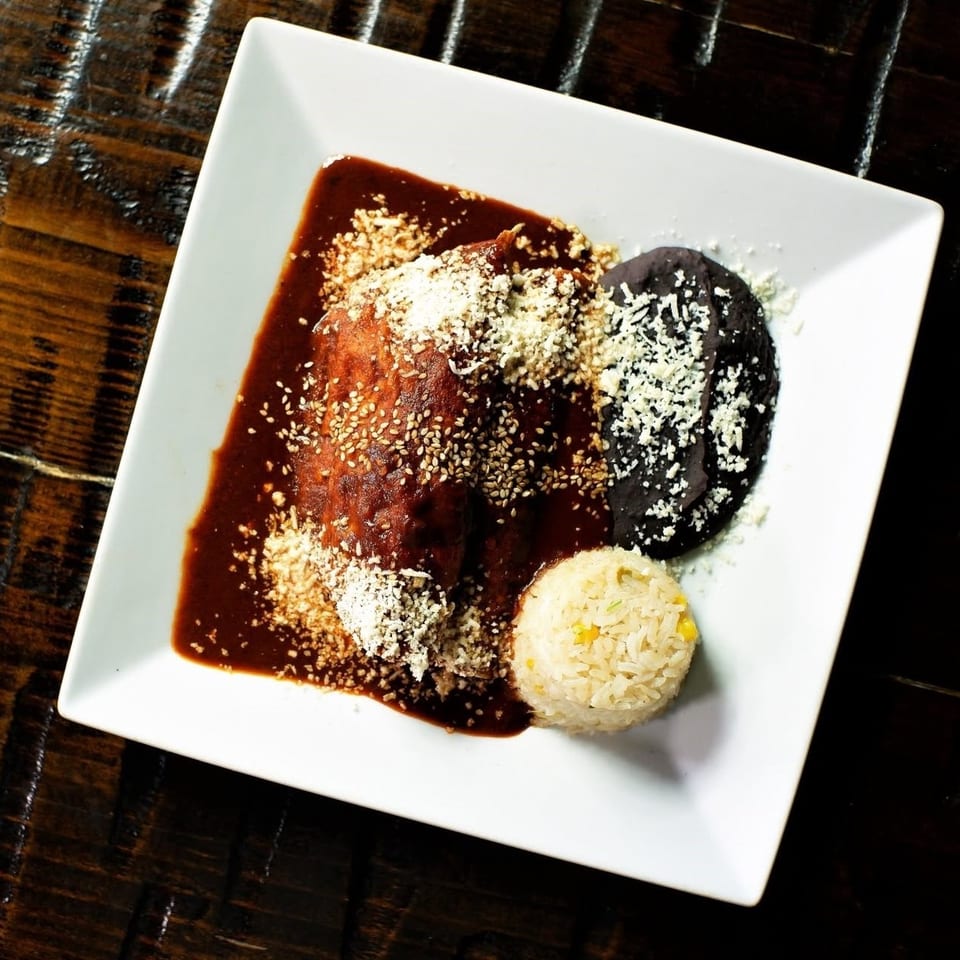 Mexican enchiladas covered in red and black mole sauces, sprinkled with sesame seeds and cheese, served with rice on a white plate.