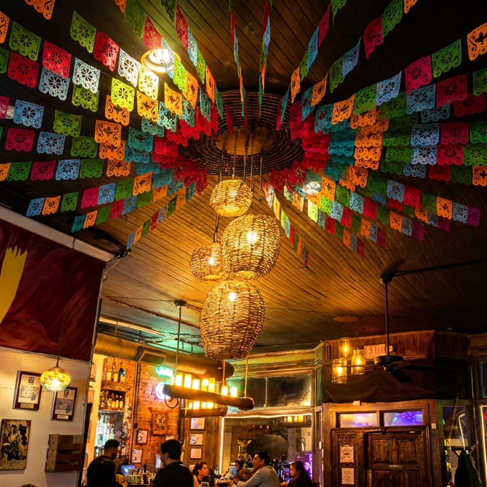 Festive Mexican restaurant interior with colorful papel picado banners, woven pendant lights, framed photographs, and diners at the bar.